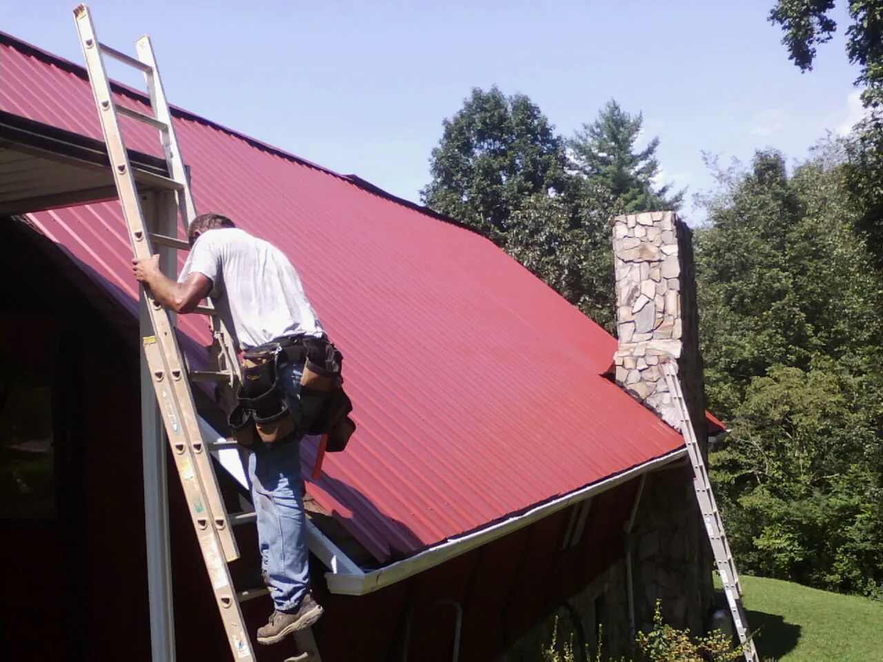 Roofer on a ladder beside a red metal roof and stone chimney — Soffit & Fascia Repair by Roofing Brothers in Maxatawny